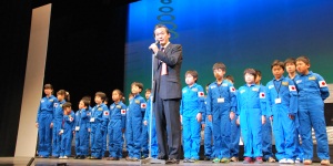 ステージで挨拶する市長と後ろに横二列に並ぶ子どもたちの写真